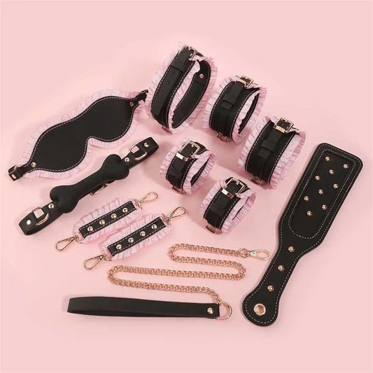 Product flatlay of the black version of the Royal Ruffle bondage set, featuring black leather with pastel pink ruffles, a studded paddle, cuffs, collar, and gag laid out on a pink background.