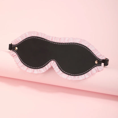 The black leather padded blindfold mask featuring a delicate pink ruffled lace trim and an adjustable strap, displayed over a pink display cylinder.