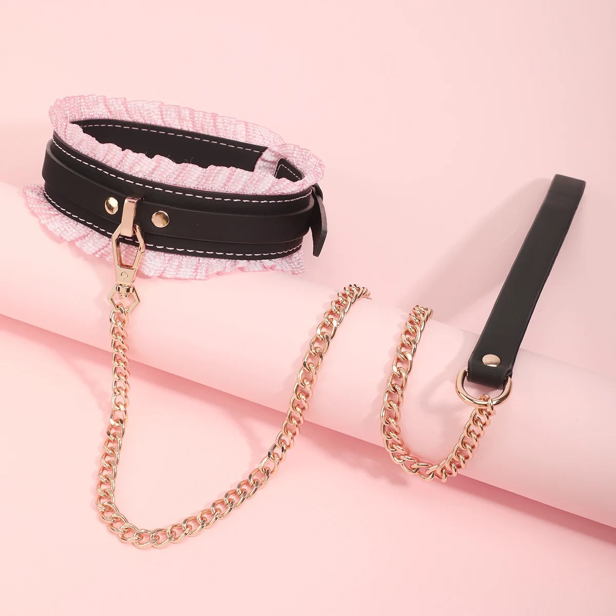 The black version of the ruffled bondage collar displayed at an angle, showing the heavy gold chain leash attached to the central D-ring over a pink prop.