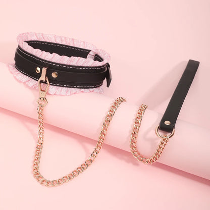 The black version of the ruffled bondage collar displayed at an angle, showing the heavy gold chain leash attached to the central D-ring over a pink prop.