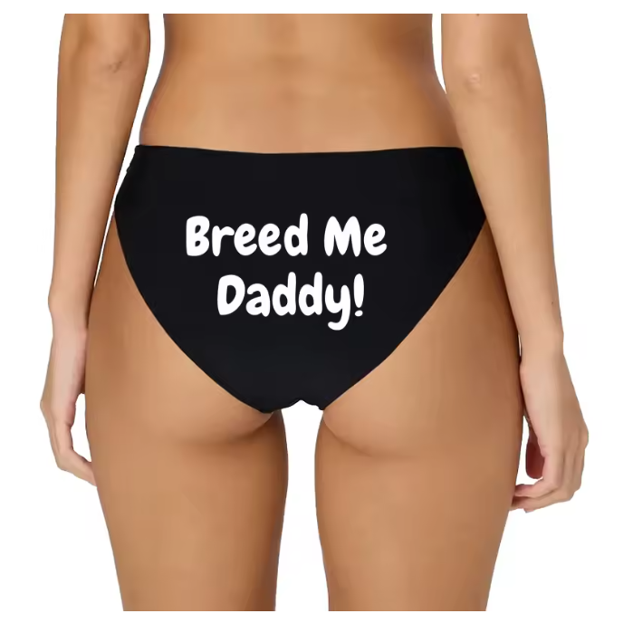 Model posing in black bikini panties with white 'Breed Me Daddy!' slogan. Kinky breeding fantasy lingerie.