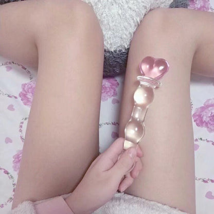 Lifestyle aesthetic shot of the pink Heart Wand resting playfully between a model's thighs on soft floral bedding.