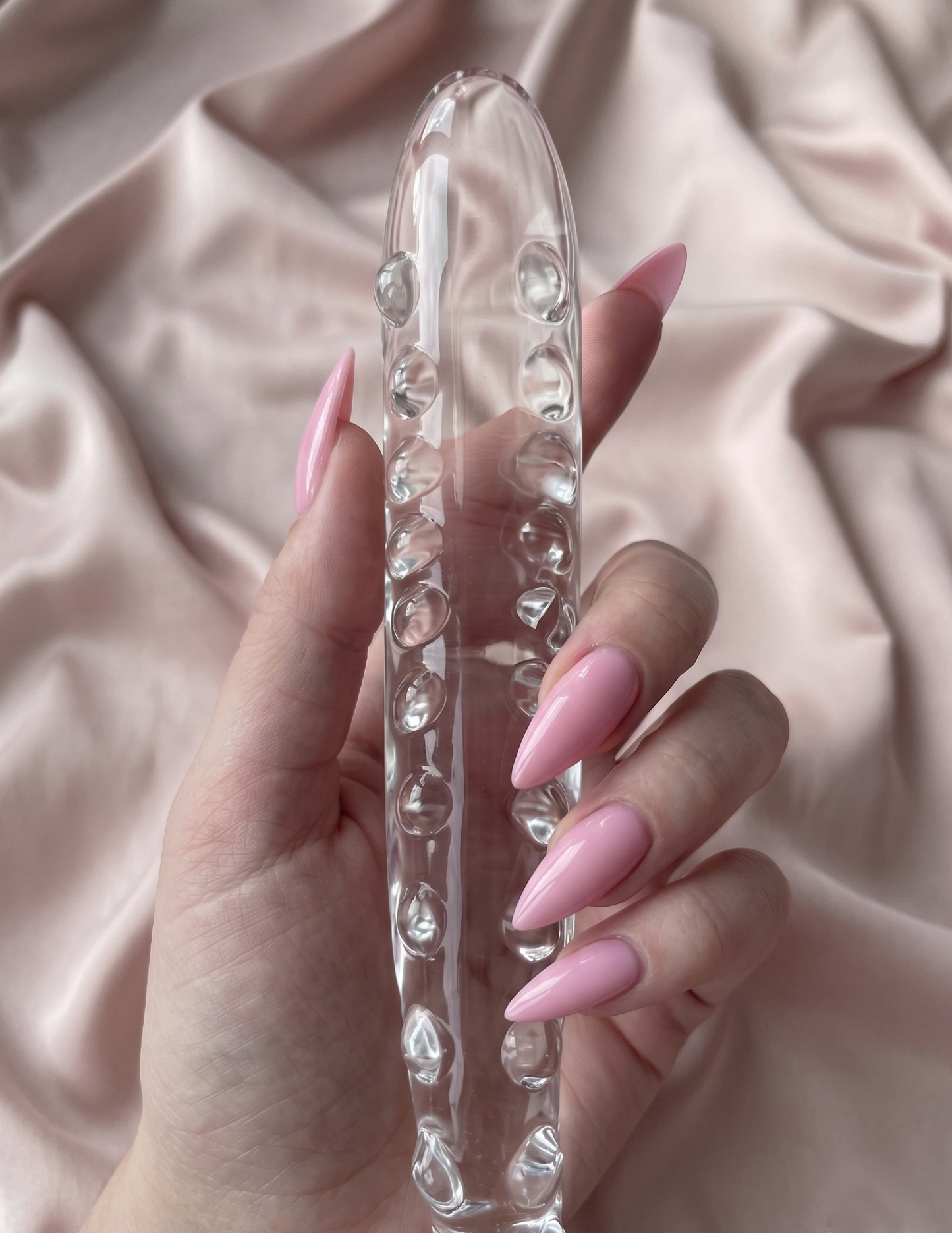 Hand with pink nails holding a crystal clear slim wave glass tentacle toy against a soft, draped silk background.