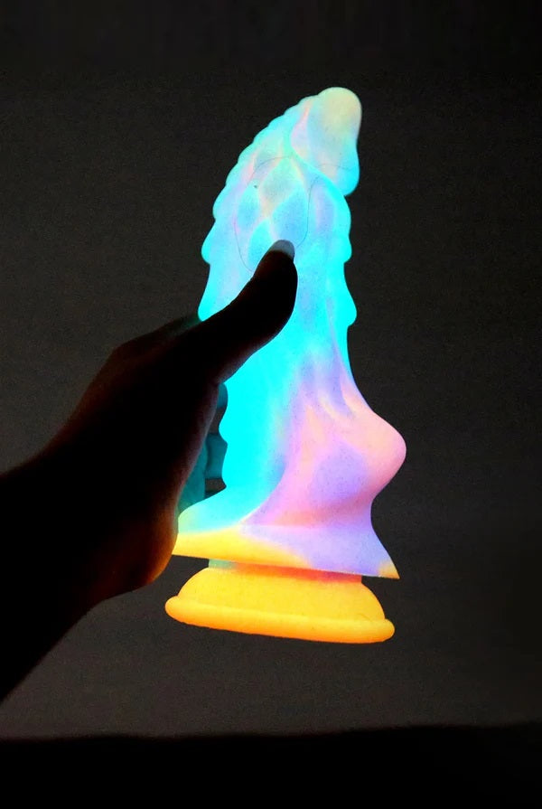 Silhouette of a hand holding the fantasy dragon dildo as it powerfully glows in the dark against a black background.