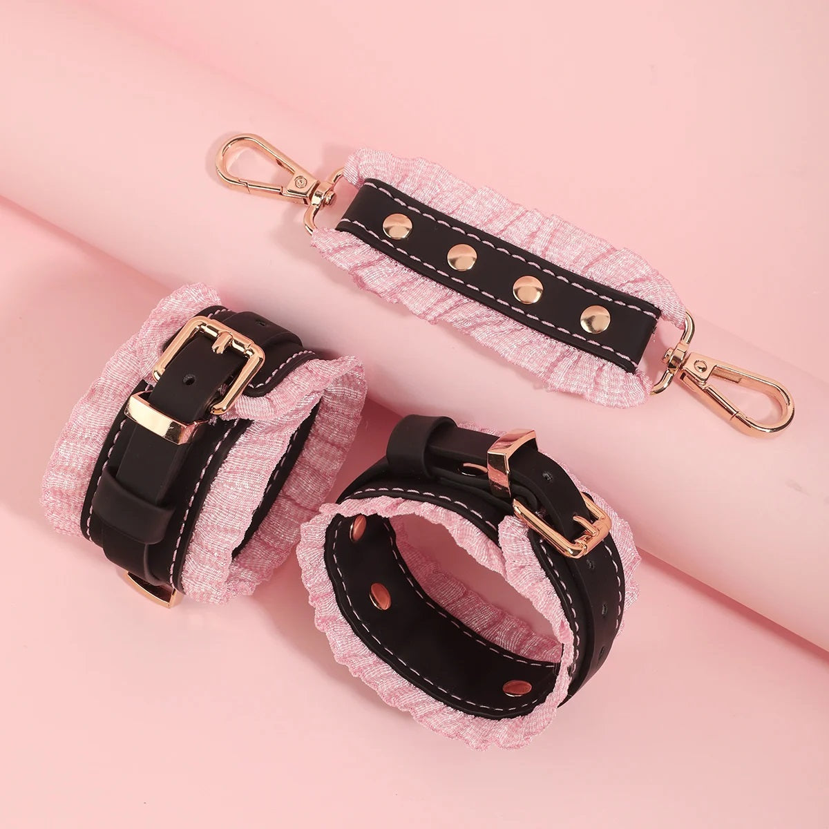 Close-up shot of the black leather cuffs featuring gold studs, pastel pink ruffled borders, and a matching black connector strap against a pink background.