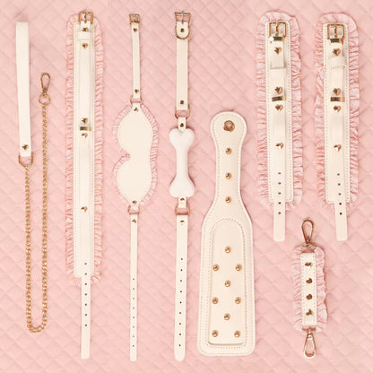 A clean grid lineup of all pieces in the white and pink bondage collection, displaying the paddle, straps, blindfold, and collars laid out perfectly on a blush pink background.