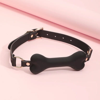 The black leather bone-shaped mouth gag with an adjustable strap and pink ruffle backing, displayed on a pastel pink background.