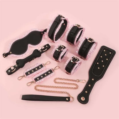 Product flatlay of the black version of the Royal Ruffle bondage set, featuring black leather with pastel pink ruffles, a studded paddle, cuffs, collar, and gag laid out on a pink background.