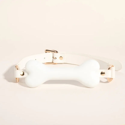 The white vegan leather bone gag with gold buckles and a pastel pink ruffled fabric lining, centered on a soft beige background.