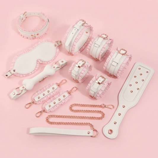Main product flatlay of the Royal Ruffle 6-piece bondage set in white vegan leather and pastel pink ruffles with rose gold hardware, including handcuffs, ankle cuffs, collar, leash, blindfold, paddle, and bone gag on a pink background.