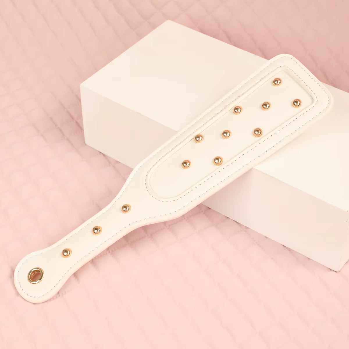 Close-up of the elegant white vegan leather impact paddle, featuring decorative rose gold studs and a comfortable curved handle resting on a white geometric block.