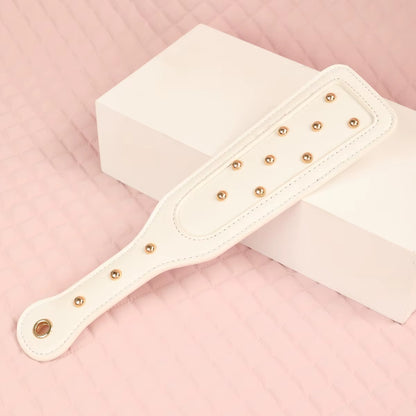 Close-up of the elegant white vegan leather impact paddle, featuring decorative rose gold studs and a comfortable curved handle resting on a white geometric block.