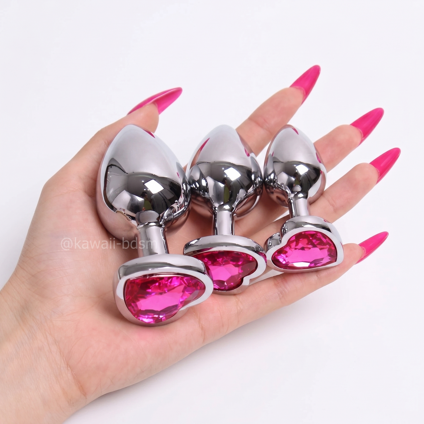 A hand with pink acrylic nails holding a 3-piece pastel pink crystal heart stainless steel plug training set.