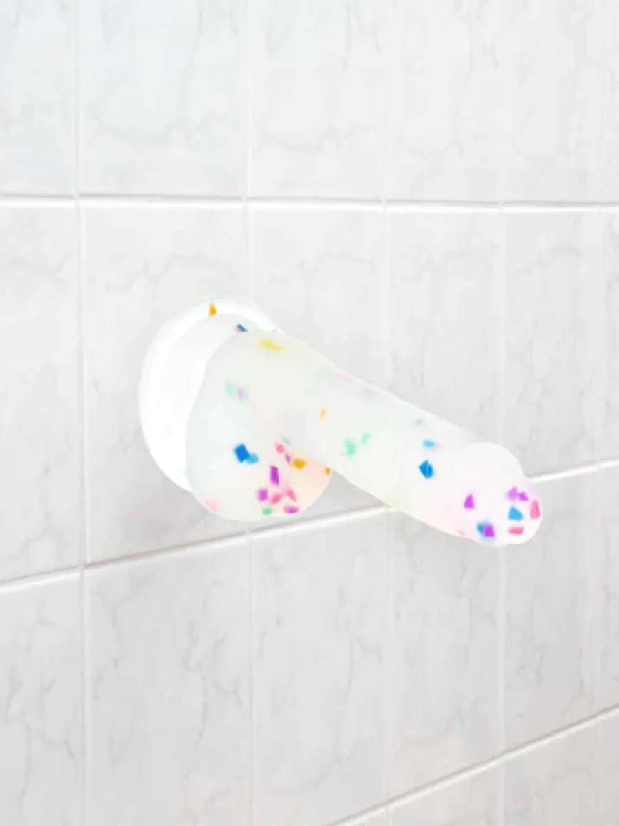 The kawaii confetti silicone dildo firmly attached to white shower tiles, demonstrating the strong suction cup for hands-free play.