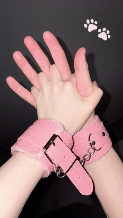 A close-up of two hands gently held together, wearing pastel pink faux leather restraint cuffs with soft fluffy lining against a dark background.