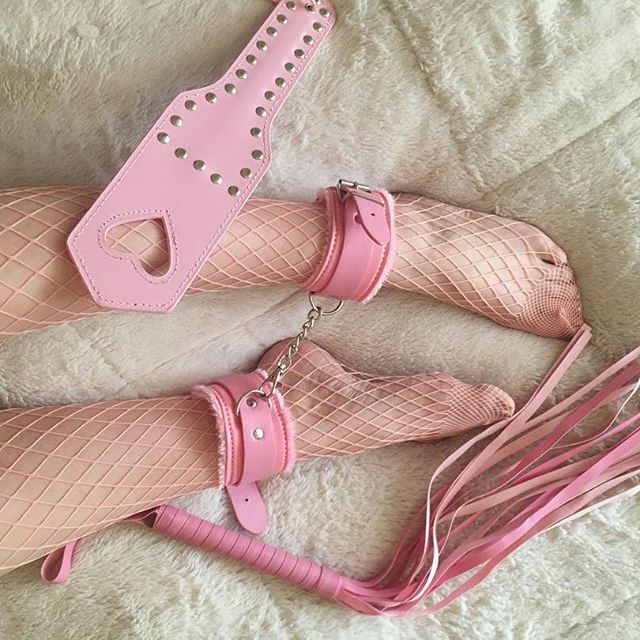 Pastel pink heart spanking paddle styled aesthetically on a bed next to pink fluffy handcuffs and bondage rope.