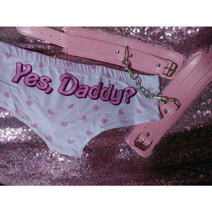 A flatlay photograph of pink "Yes, Daddy?" heart-print panties, surrounded by cute pink handcuffs