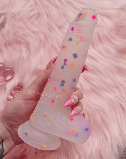Hand holding the cute translucent confetti dildo with colorful sprinkles against an aesthetic soft pink faux fur background.