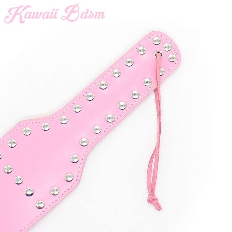 Angled view of the cute pink spanking paddle showcasing the shiny silver studs and the attached security wrist strap.